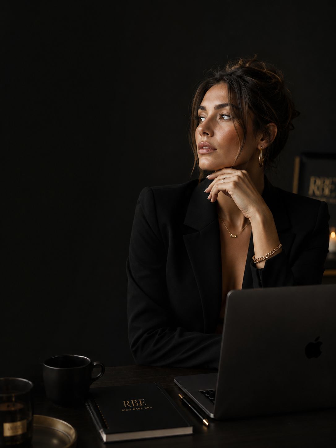A Rich Babe Era creator seated at her desk in soft low light, laptop open beside a gold-embossed RBE notebook