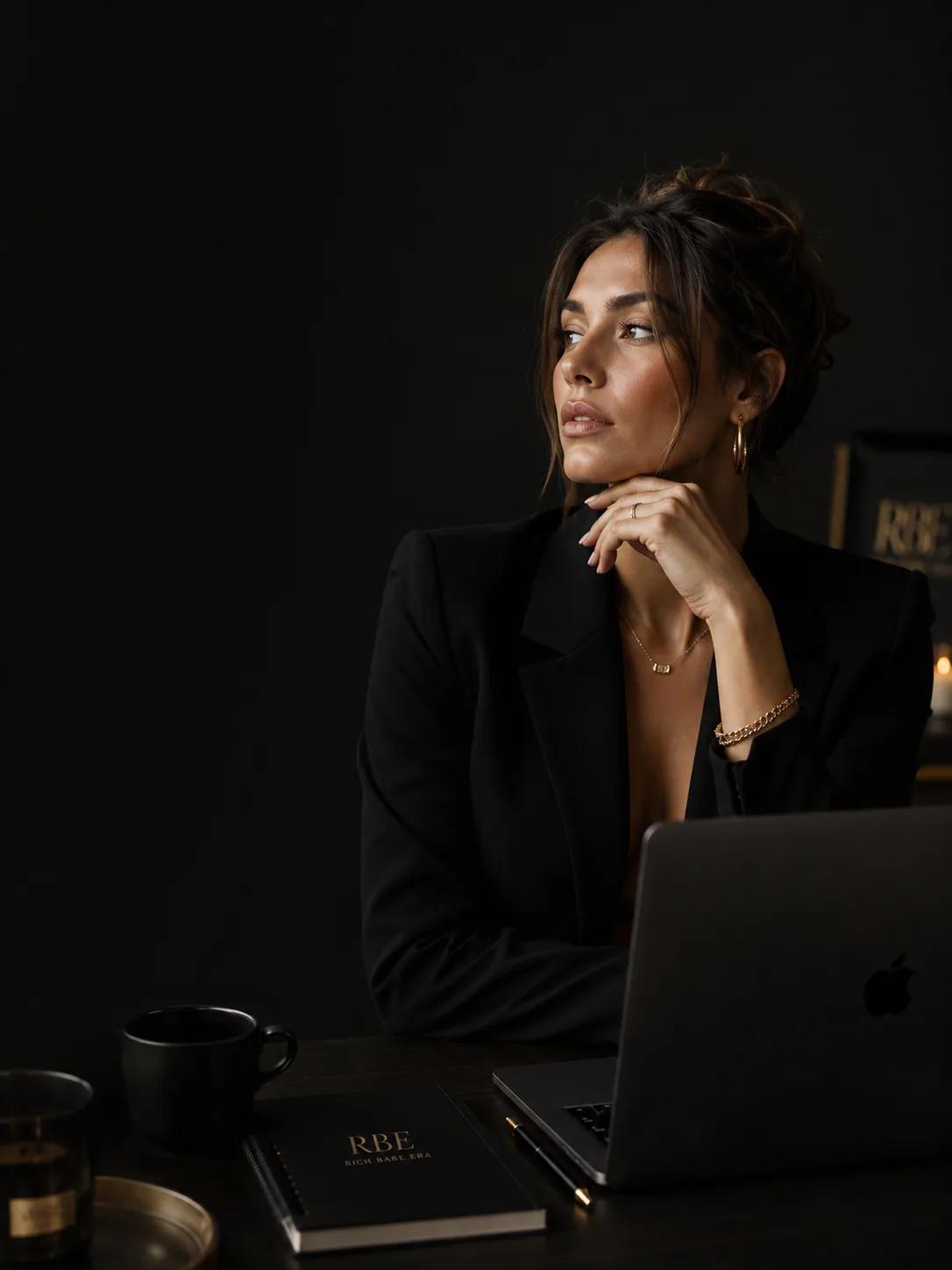A Rich Babe Era creator seated at her desk in soft low light, laptop open beside a gold-embossed RBE notebook