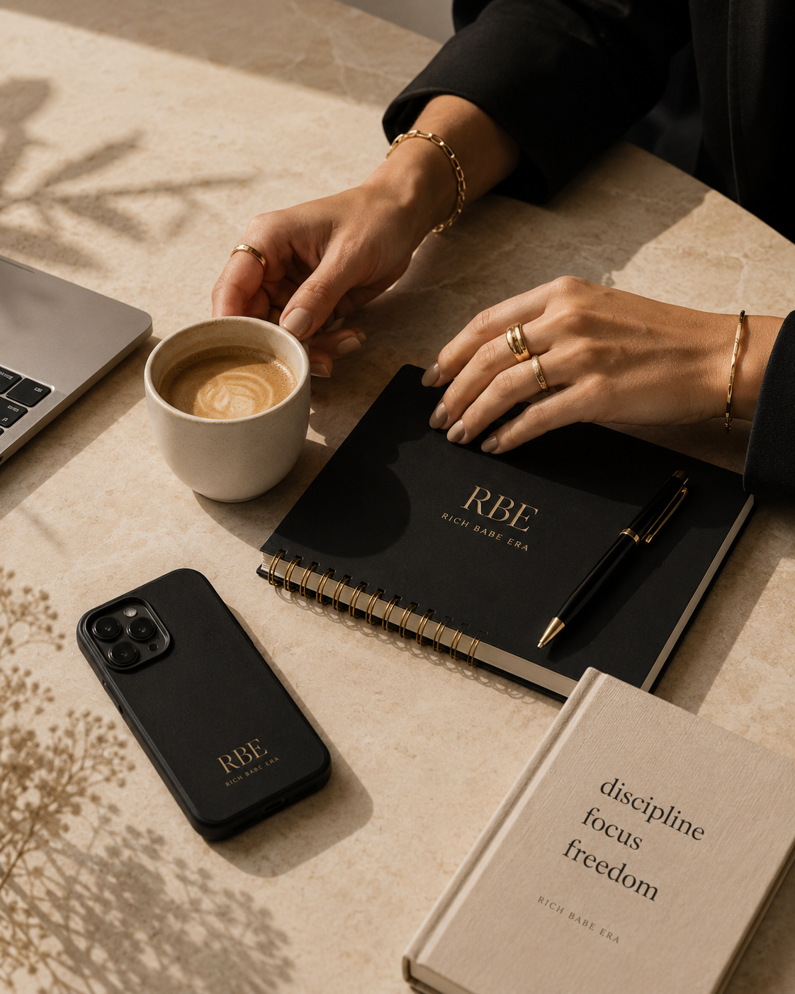 A woman’s hands with gold jewellery beside a black RBE notebook, phone and coffee on a warm cream stone surface