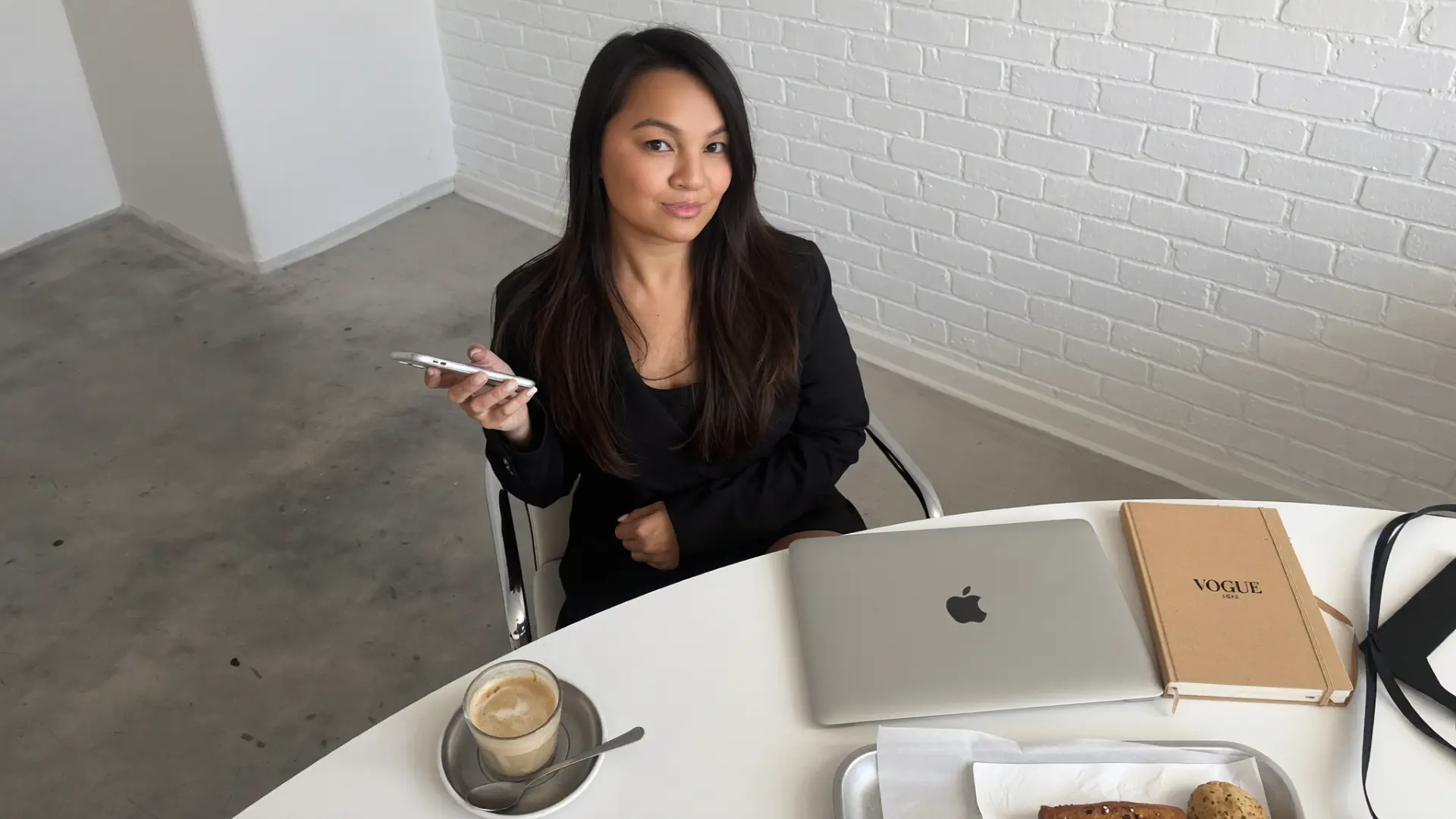 Emily, the founder of Rich Babe Era, seated at a white table with her laptop, phone, coffee and a Vogue notebook